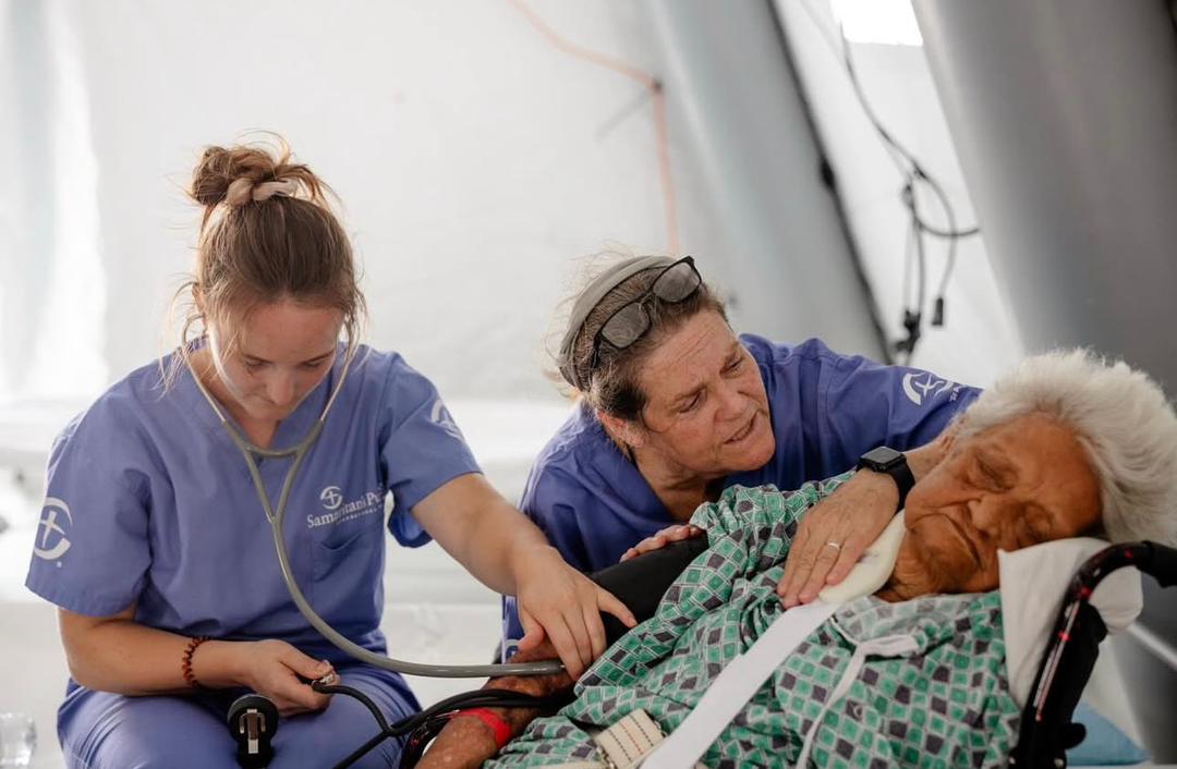 Two nurses in an emergency field hospital attending to an elderly woman resting in a wheelchair